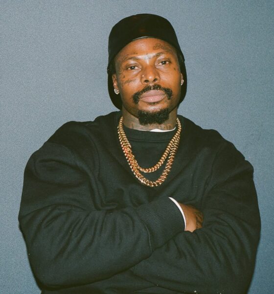 A studio portrait of Asake wearing a black sweatshirt, two gold Cuban link chains, and a black beanie, posing with his arms crossed against a neutral grey background.
