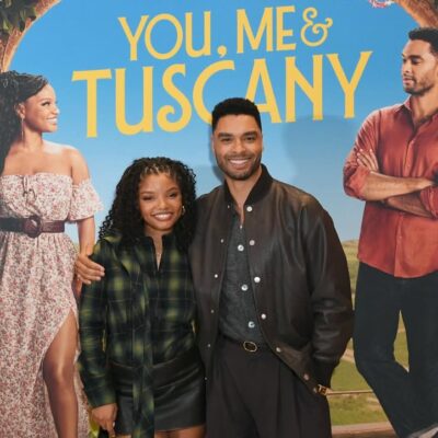 Halle Bailey and Regé-Jean Page posing together in front of a "You, Me & Tuscany" movie poster featuring them in a romantic Italian setting.