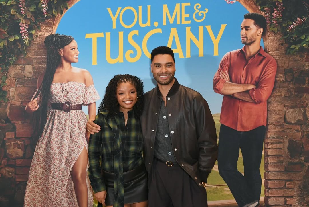 Halle Bailey and Regé-Jean Page posing together in front of a "You, Me & Tuscany" movie poster featuring them in a romantic Italian setting.