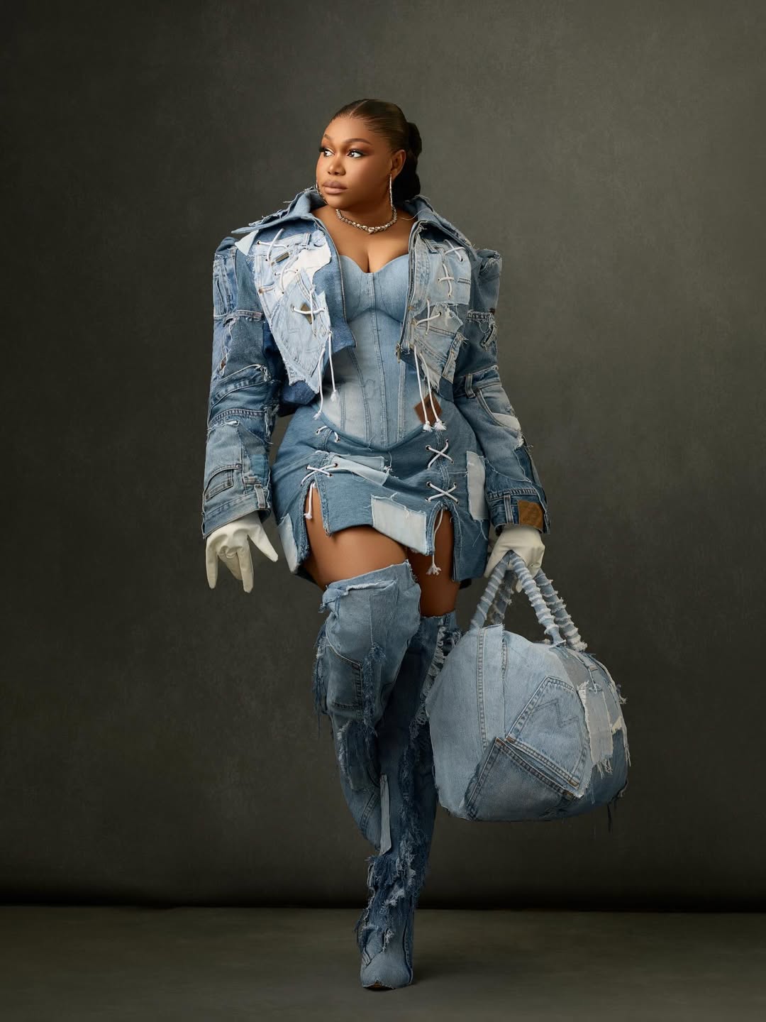 Nollywood actress Ruth Kadiri standing and posing in a deconstructed upcycled denim corset top and mini-skirt, paired with dramatic thigh-high denim boots. She wears a single white glove and has a distressed denim duffle bag on the floor beside her.