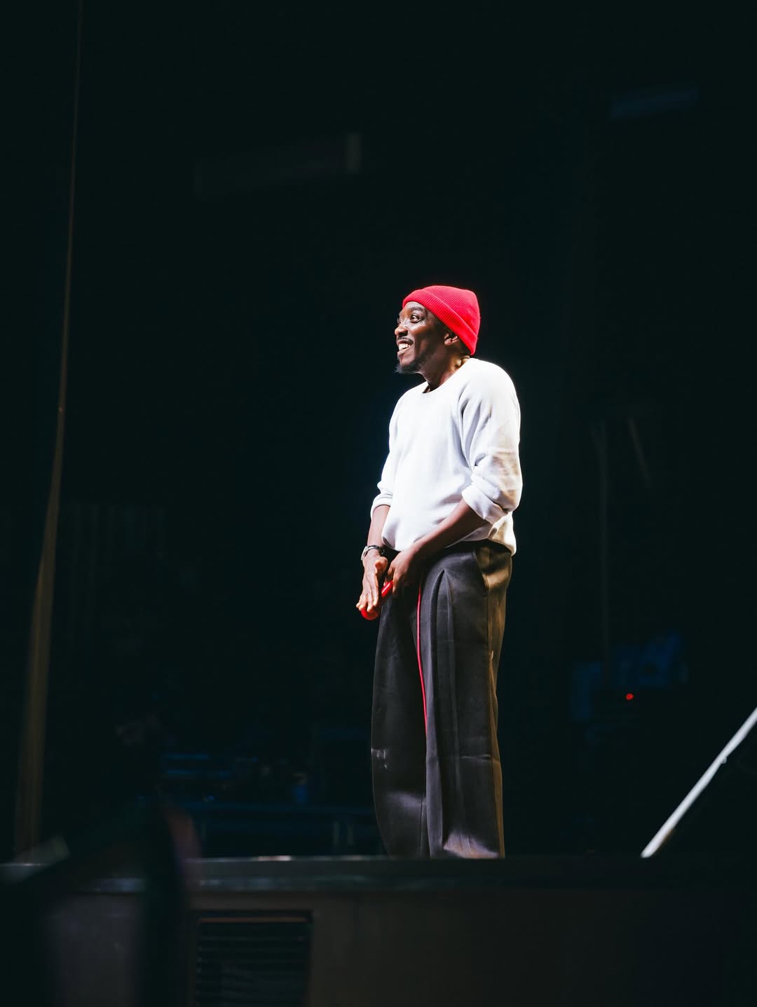 Nigerian comedian Bovi Ugboma performing on stage in a white sweatshirt and red beanie during his "Colour Me Blacker" comedy show in London.