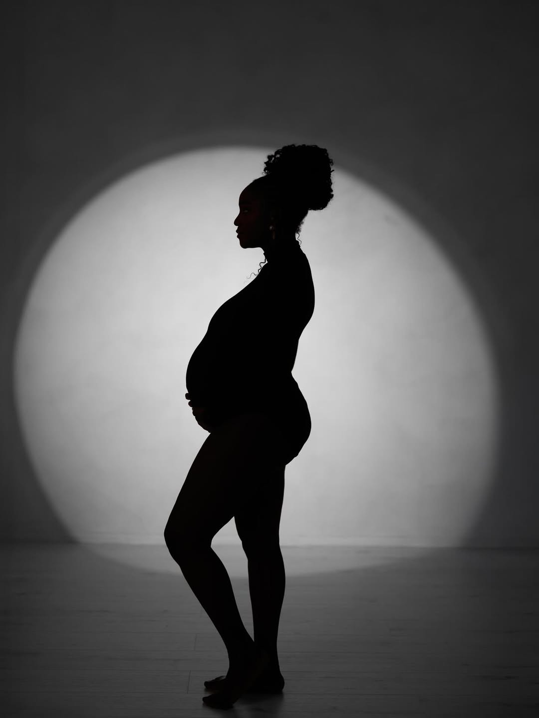 A black-and-white profile silhouette of singer Simi standing against a circular backlit glow. She is wearing a form-fitting bodysuit that highlights her pregnancy bump, with her hair styled in a high curly updo.