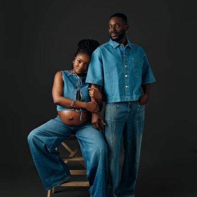 Singer Simi and her husband Adekunle Gold posing in matching mid-wash denim outfits. Simi is seated, wearing a lace-up denim crop top and wide-leg jeans, showing her baby bump. Adekunle stands beside her in a short-sleeved denim button-down and straight-leg jeans.