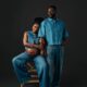 Singer Simi and her husband Adekunle Gold posing in matching mid-wash denim outfits. Simi is seated, wearing a lace-up denim crop top and wide-leg jeans, showing her baby bump. Adekunle stands beside her in a short-sleeved denim button-down and straight-leg jeans.