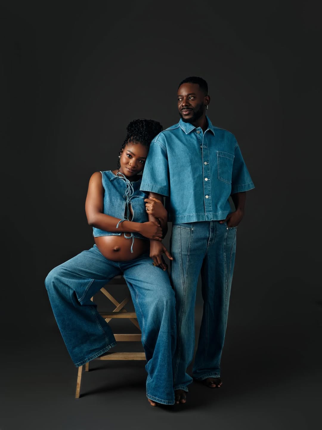 Singer Simi and her husband Adekunle Gold posing in matching mid-wash denim outfits. Simi is seated, wearing a lace-up denim crop top and wide-leg jeans, showing her baby bump. Adekunle stands beside her in a short-sleeved denim button-down and straight-leg jeans.