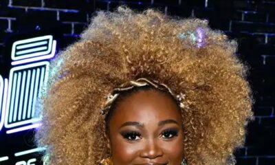 A portrait of Alexia Jayy smiling in a metallic gold fringed dress with a beaded neckline in front of The Voice "Battle of the Champions" signage.