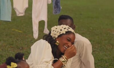 Wofai Fada laughing during an outdoor picnic pregnancy reveal. She is wearing a cream puff-sleeve top and floral headpiece, sitting on a white blanket with Taiwo Cole. Their daughter, Ifedayo, is seen in the foreground by a basket of sunflowers.