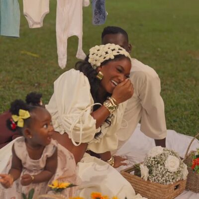 Wofai Fada laughing during an outdoor picnic pregnancy reveal. She is wearing a cream puff-sleeve top and floral headpiece, sitting on a white blanket with Taiwo Cole. Their daughter, Ifedayo, is seen in the foreground by a basket of sunflowers.