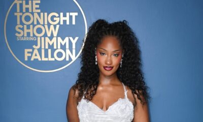 A professional portrait of Tems standing against a blue wall featuring The Tonight Show Starring Jimmy Fallon logo, wearing a silver mesh and lace gown.