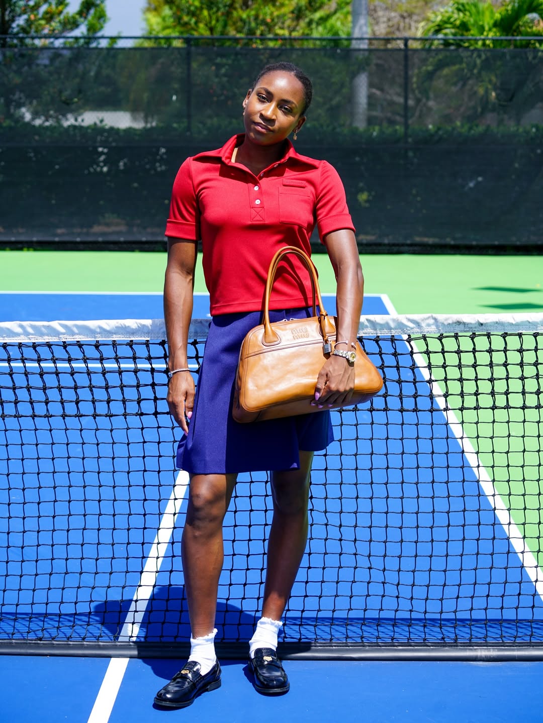 Coco Gauff posing on a tennis court in a red Miu Miu polo, navy wrap skirt, and black loafers with frilly socks for MiuMiuSS26.