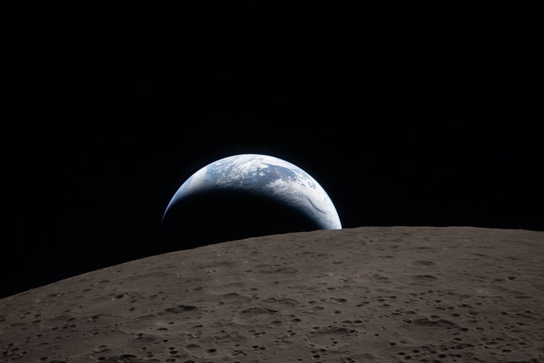 A muted blue Earth with white clouds setting behind the cratered lunar surface and Ohm crater during the Artemis II mission.