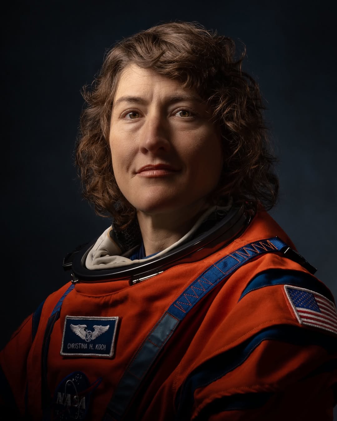 Astronaut Christina Hammock Koch posing in an orange Artemis flight suit with mission patches and a U.S. flag.