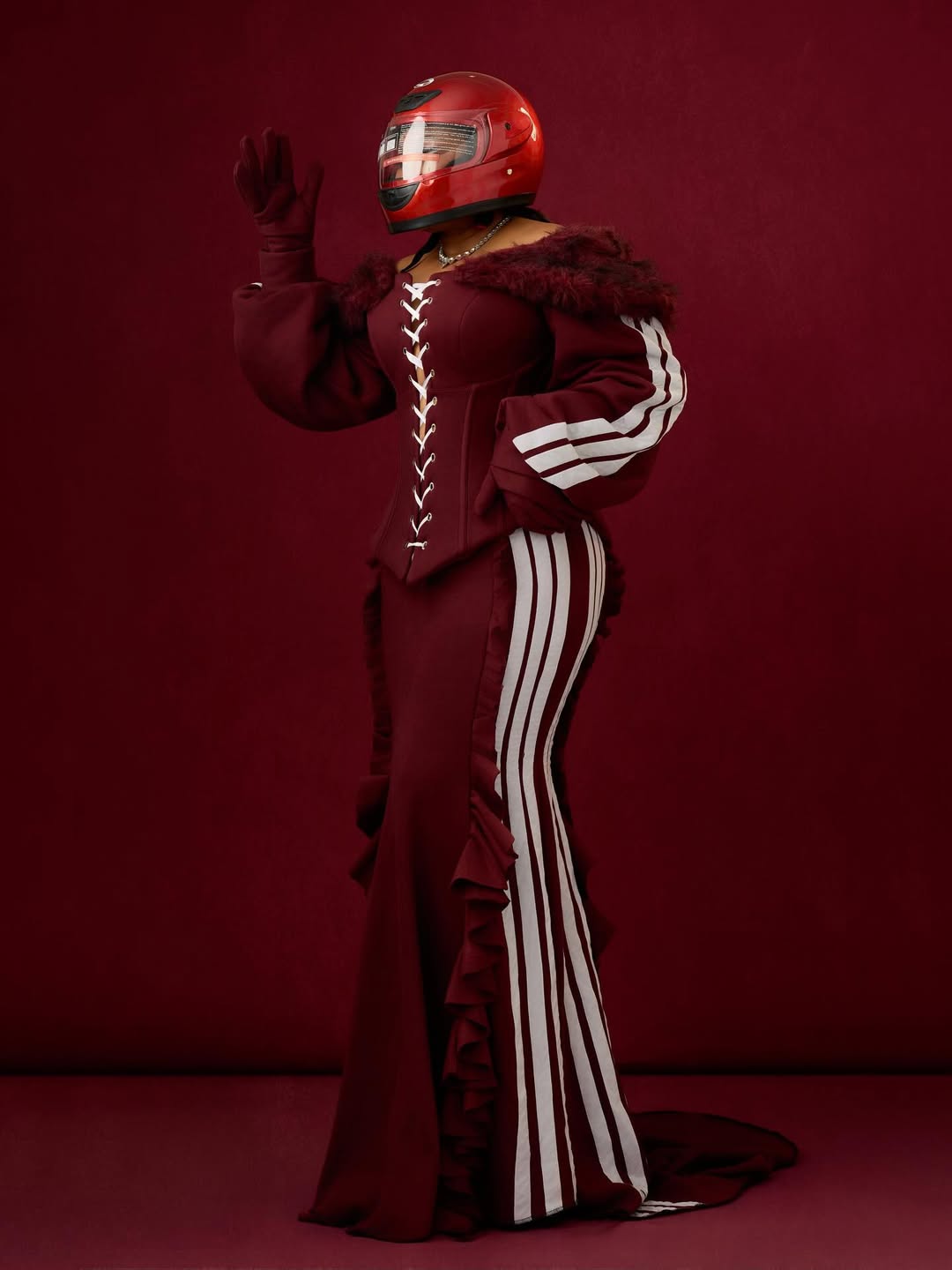 Nollywood actress Ruth Kadiri posing against a red background, wearing a high-shine red racing helmet. She is dressed in a custom burgundy two-piece set by Josiphiah Michael, featuring a structural, lace-up corset top with white racing stripes and a matching ruffled maxi skirt with off-the-shoulder fur trim.