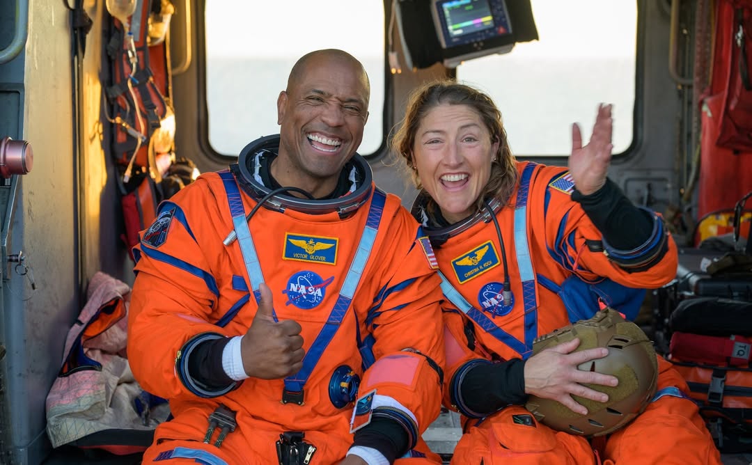 NASA astronauts Victor Glover and Christina Koch smiling inside a Navy MH-60 Seahawk helicopter on the flight deck of the USS John P. Murtha.