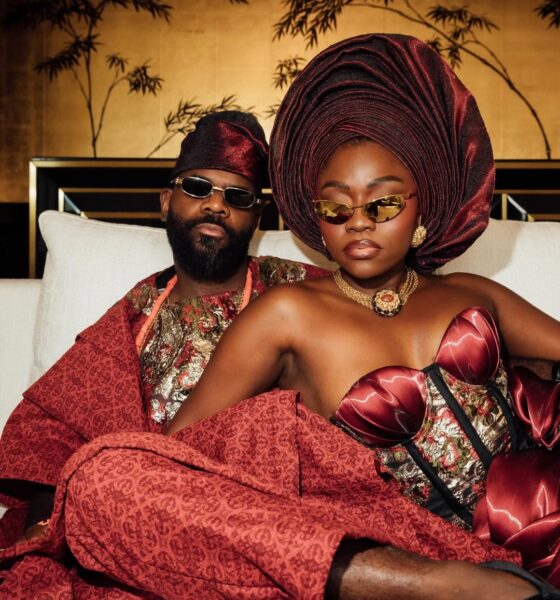 Fisayo Longe in a strapless crimson and black brocade gown with a structural bow, standing with Afolabi Mosuro in a matching Agbada in front of a white wall.