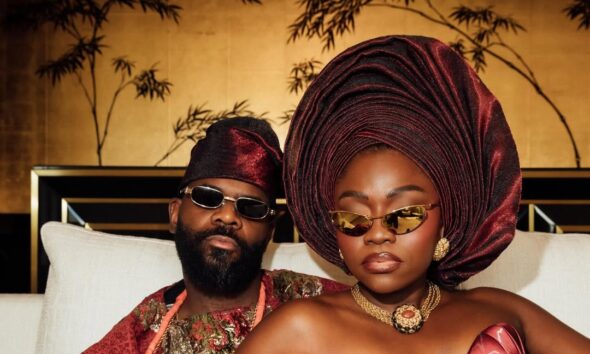 Fisayo Longe in a strapless crimson and black brocade gown with a structural bow, standing with Afolabi Mosuro in a matching Agbada in front of a white wall.