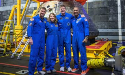 The four Artemis II astronauts—Reid Wiseman, Christina Koch, Jeremy Hansen, and Victor Glover—pose in blue flight suits in front of the Orion capsule in the well deck of the USS John P. Murtha.