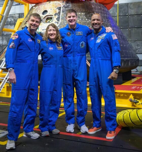 The four Artemis II astronauts—Reid Wiseman, Christina Koch, Jeremy Hansen, and Victor Glover—pose in blue flight suits in front of the Orion capsule in the well deck of the USS John P. Murtha.