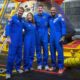 The four Artemis II astronauts—Reid Wiseman, Christina Koch, Jeremy Hansen, and Victor Glover—pose in blue flight suits in front of the Orion capsule in the well deck of the USS John P. Murtha.