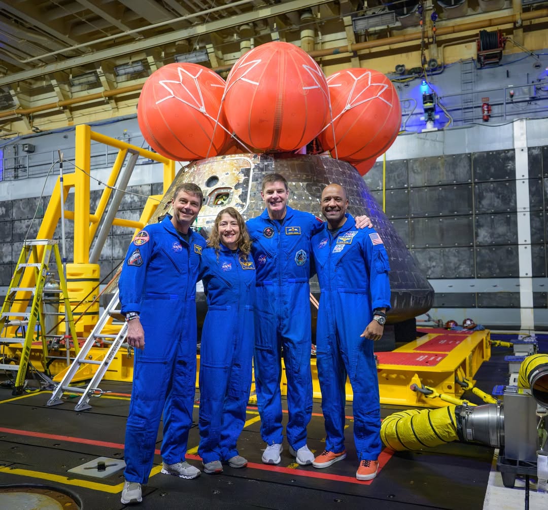 The four Artemis II astronauts—Reid Wiseman, Christina Koch, Jeremy Hansen, and Victor Glover—pose in blue flight suits in front of the Orion capsule in the well deck of the USS John P. Murtha.