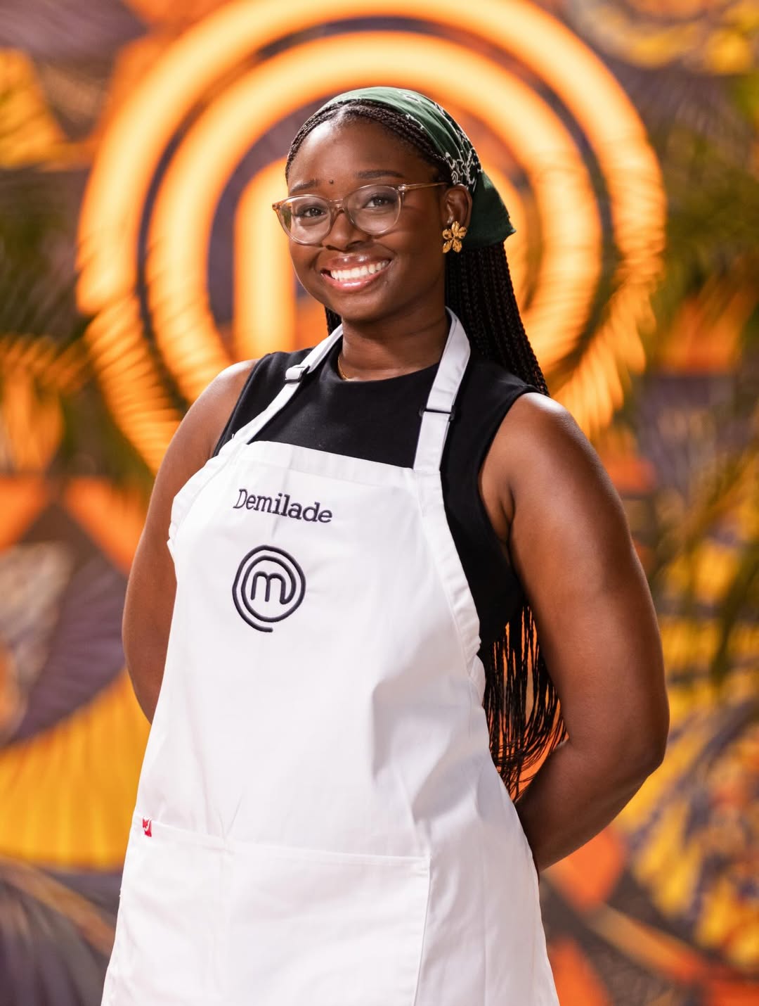 MasterChef Nigeria contestant Demilade Akingbe smiling in her white apron, wearing glasses and a green headscarf.