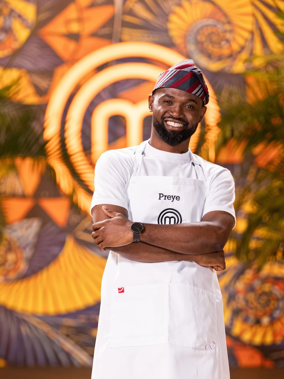MasterChef Nigeria contestant Preye Fadese smiling with arms crossed, wearing a white apron and a traditional striped red and black cap.