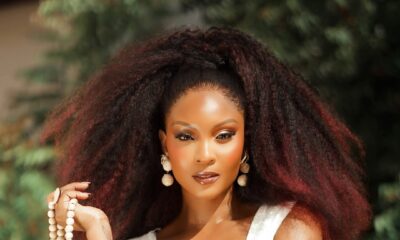 Osas Ighodaro with voluminous, red-tinted kinky-coily hair, wearing a white textured crop top and fringe skirt with heavy gold bangles and beaded accessories.