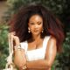 Osas Ighodaro with voluminous, red-tinted kinky-coily hair, wearing a white textured crop top and fringe skirt with heavy gold bangles and beaded accessories.