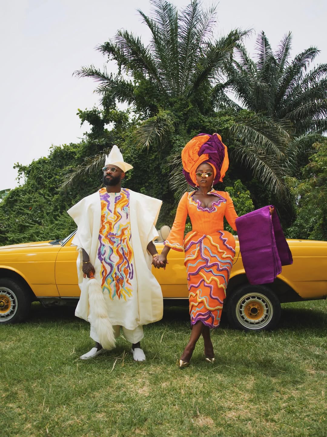 Fisayo Longe and Afolabi Mosuro holding hands in front of a yellow vintage convertible car wearing orange and purple embroidered traditional attire.
