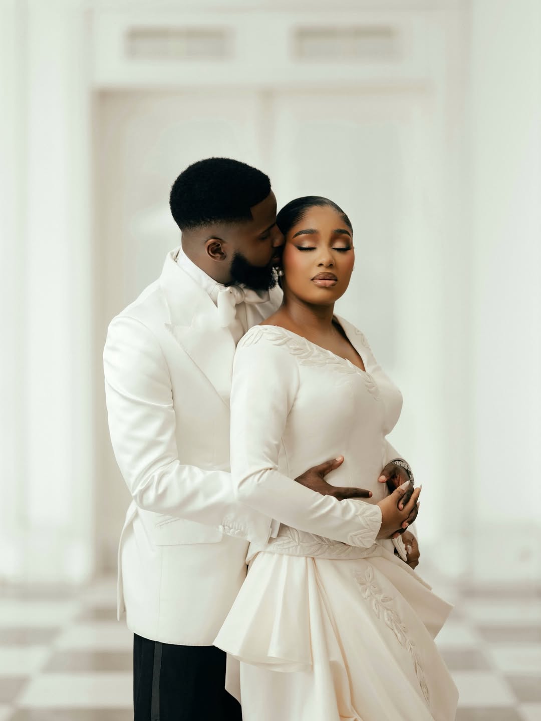 An intimate portrait of Akin Faminu embracing Kiky Festus from behind. Akin wears a white Deji and Kola dinner jacket; Kiky wears a bespoke ivory Mazelle Bridal two-piece set.