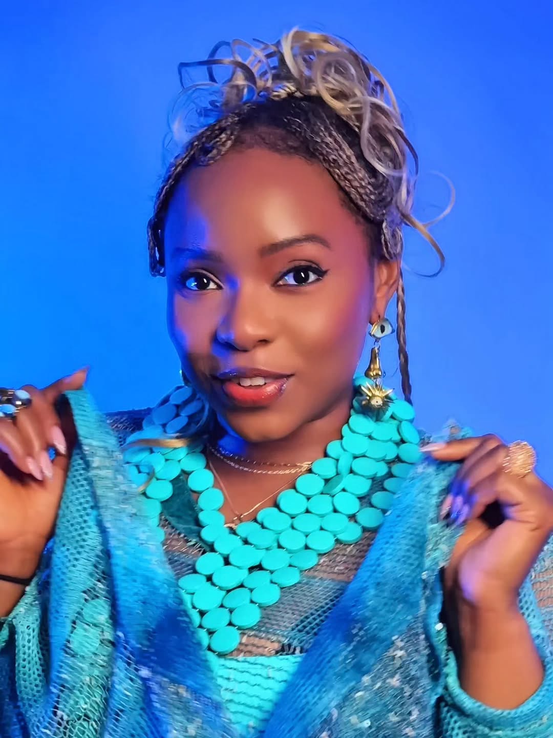 Close-up of Yemi Alade wearing a heavy turquoise beaded necklace and a blue mesh outfit with a blonde-streaked braided updo.