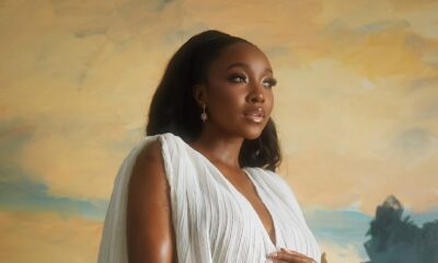 Nollywood actor Ini Dima-Okojie posing in a white pleated maternity gown with dramatic cape sleeves against an artistic painted backdrop.