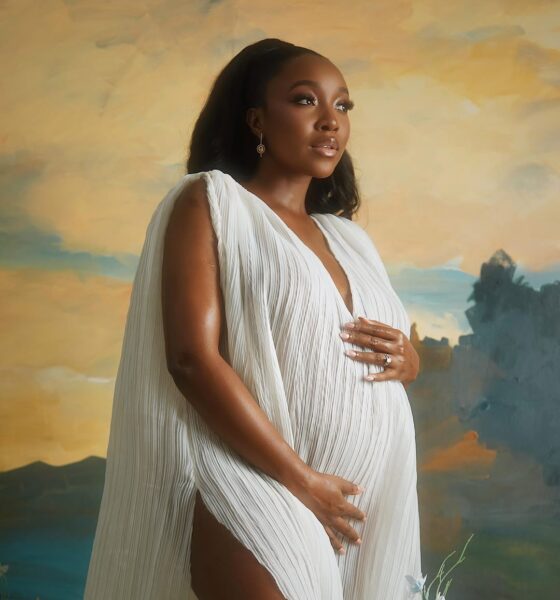 Nollywood actor Ini Dima-Okojie posing in a white pleated maternity gown with dramatic cape sleeves against an artistic painted backdrop.
