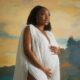 Nollywood actor Ini Dima-Okojie posing in a white pleated maternity gown with dramatic cape sleeves against an artistic painted backdrop.