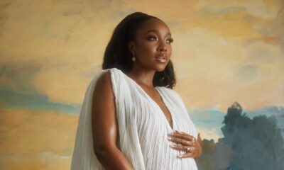 A close-up portrait of Ini Dima-Okojie holding her baby bump, wearing a sheer white V-neck maternity gown with clean, dewy makeup.