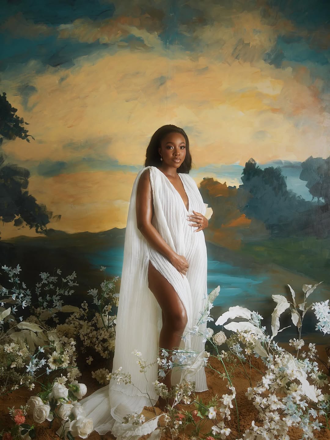 Actress Ini Dima-Okojie posing in a white pleated Toju Foyeh gown against an impressionist-style painted landscape backdrop with white roses.