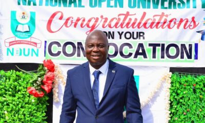 Nollywood actor Bolaji Amusan (Mr Latin) in a navy blue suit standing in front of a National Open University of Nigeria (NOUN) graduation banner.
