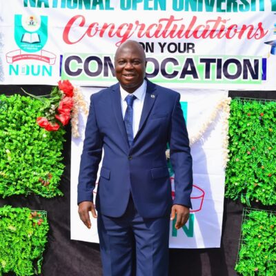 Nollywood actor Bolaji Amusan (Mr Latin) in a navy blue suit standing in front of a National Open University of Nigeria (NOUN) graduation banner.