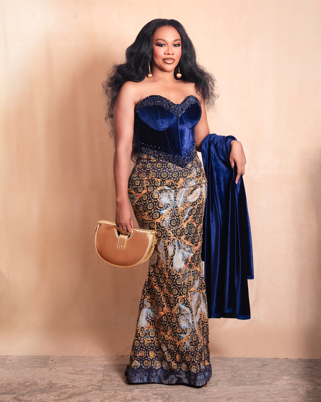 Actor Stella Damasus posing in a beaded midnight blue velvet corset and a gold patterned floor-length skirt while holding a gold clutch.