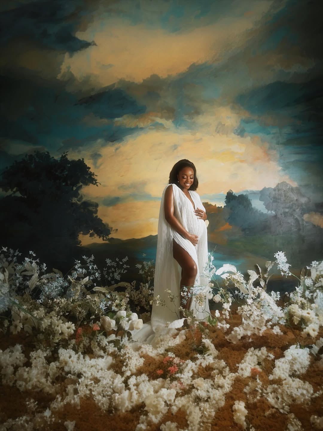 Ini Dima-Okojie in a white gown with a deep side slit, standing on a set decorated with trailing white blooms and golden-sky backdrop.