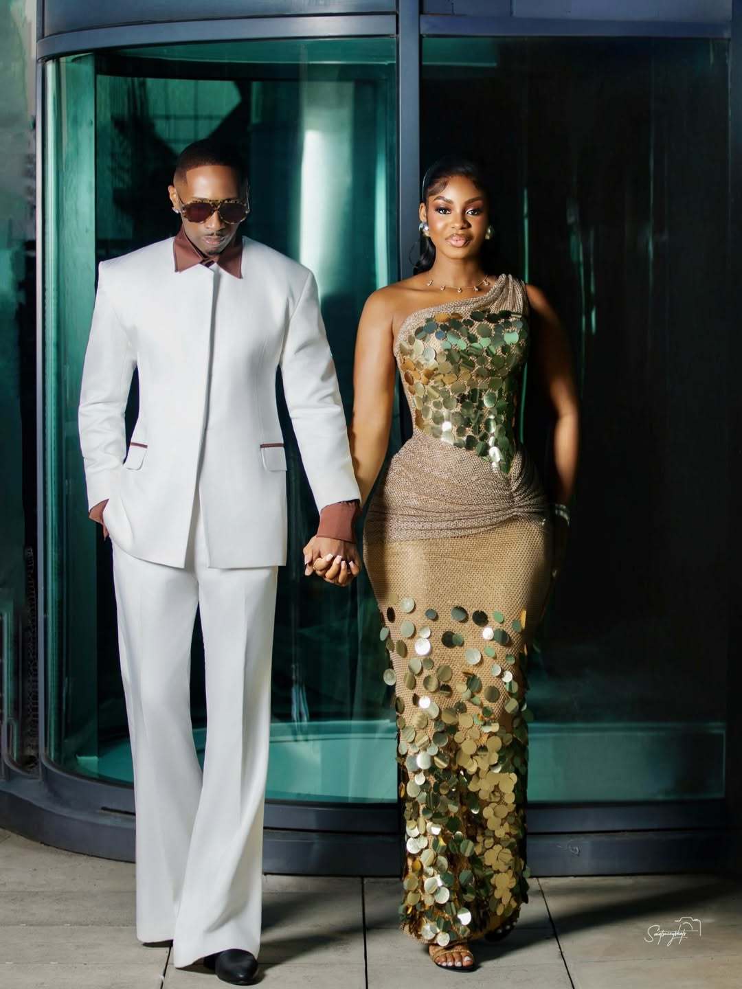 Juma Jux in a white Swazzi suit and Priscilla Ojo in a gold sequin Styled By Layo gown holding hands.