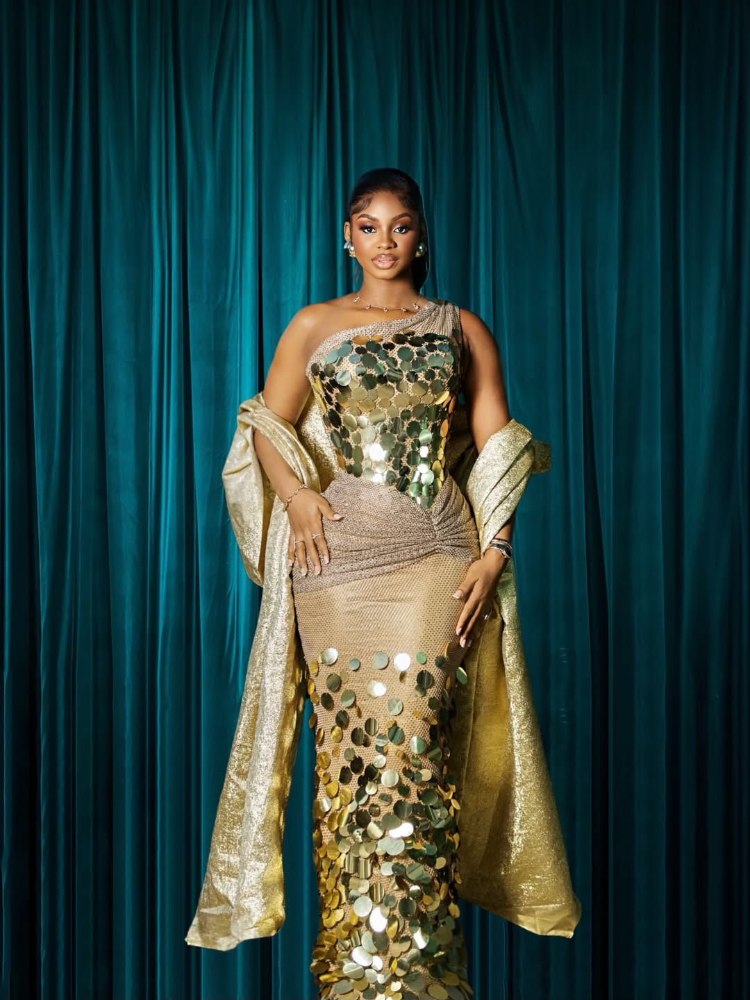 Priscilla Ojo wearing a one-shoulder gold metallic disc gown by Styled By Layo with a matching gold wrap.