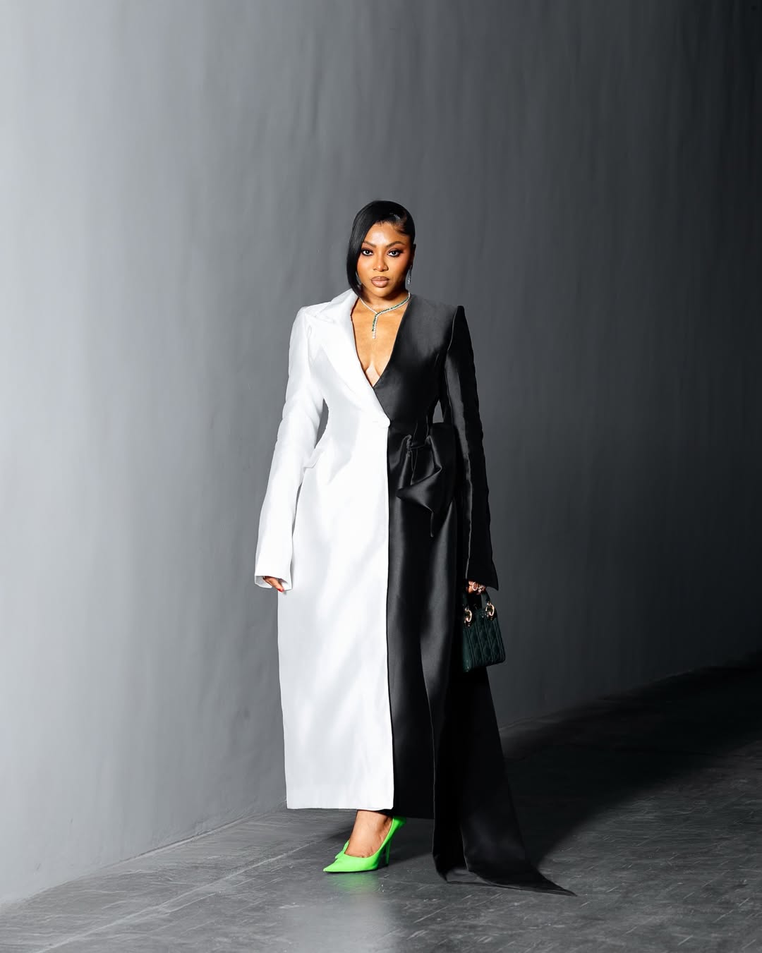 Stephanie Coker posing in a FRUCHÉ black and white split-tone coat dress with neon green heels and a dark green mini bag.