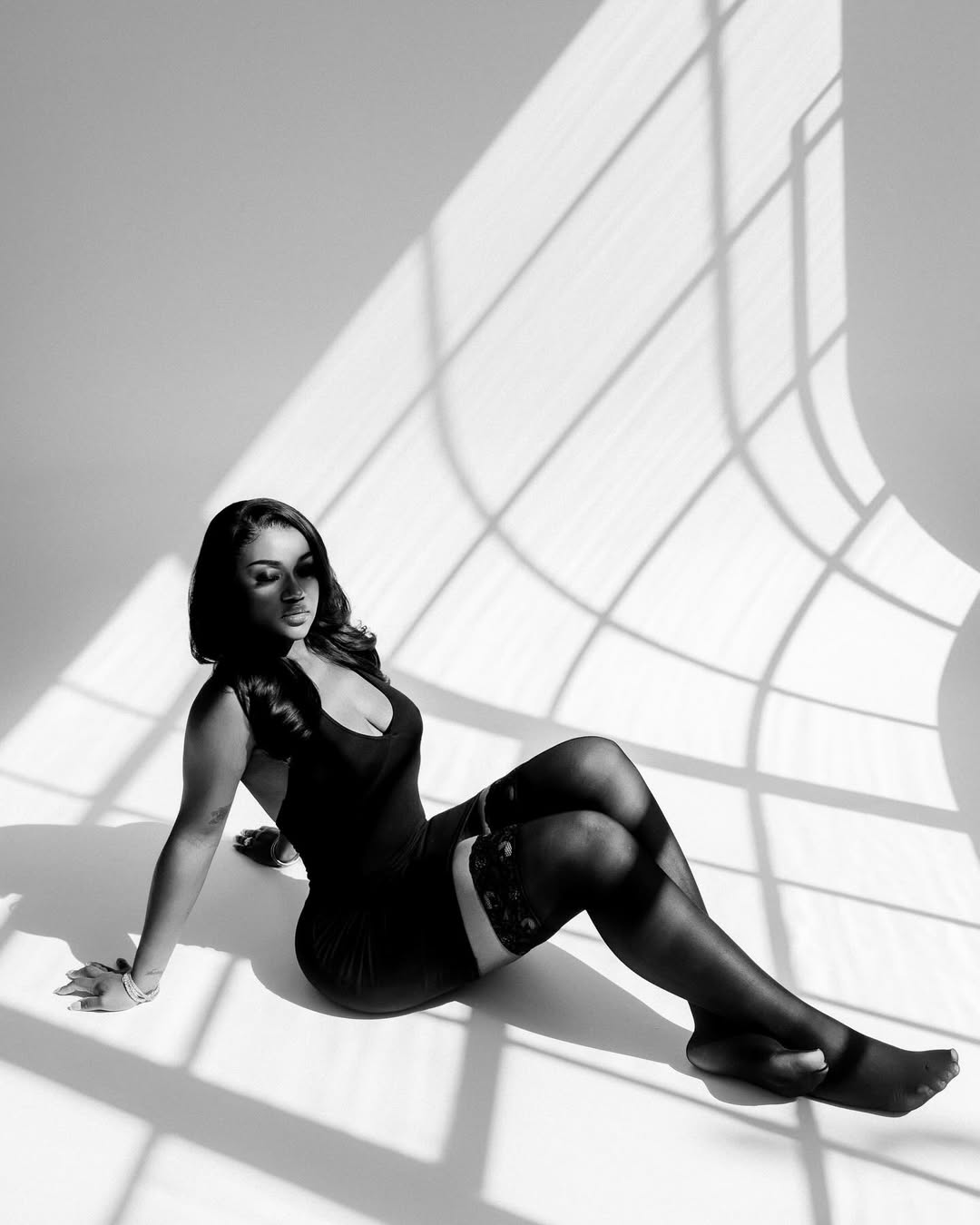 A black and white editorial shot of Chioma Adeleke sitting on the floor in a black dress and stockings.