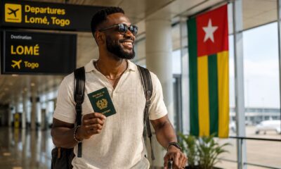 An AI-generated photo of a smiling Ghanaian traveller holding an ECOWAS Ghana passport at Lomé-Gnassingbé Eyadéma International Airport, looking towards the large Togo flag on display in the departures terminal, representing Togo's new 30-day visa-free entry for African citizens.