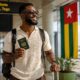 An AI-generated photo of a smiling Ghanaian traveller holding an ECOWAS Ghana passport at Lomé-Gnassingbé Eyadéma International Airport, looking towards the large Togo flag on display in the departures terminal, representing Togo's new 30-day visa-free entry for African citizens.