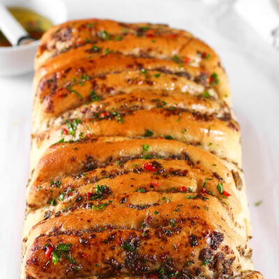 A golden-brown, freshly baked loaf of Kikifoodies' sardine pull-apart bread brushed with glossy, spicy oil and topped with chopped parsley and red pepper flakes.