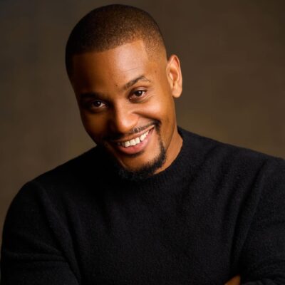 A close-up studio portrait of Nollywood actor Alexx Ekubo smiling and wearing a black jumper.