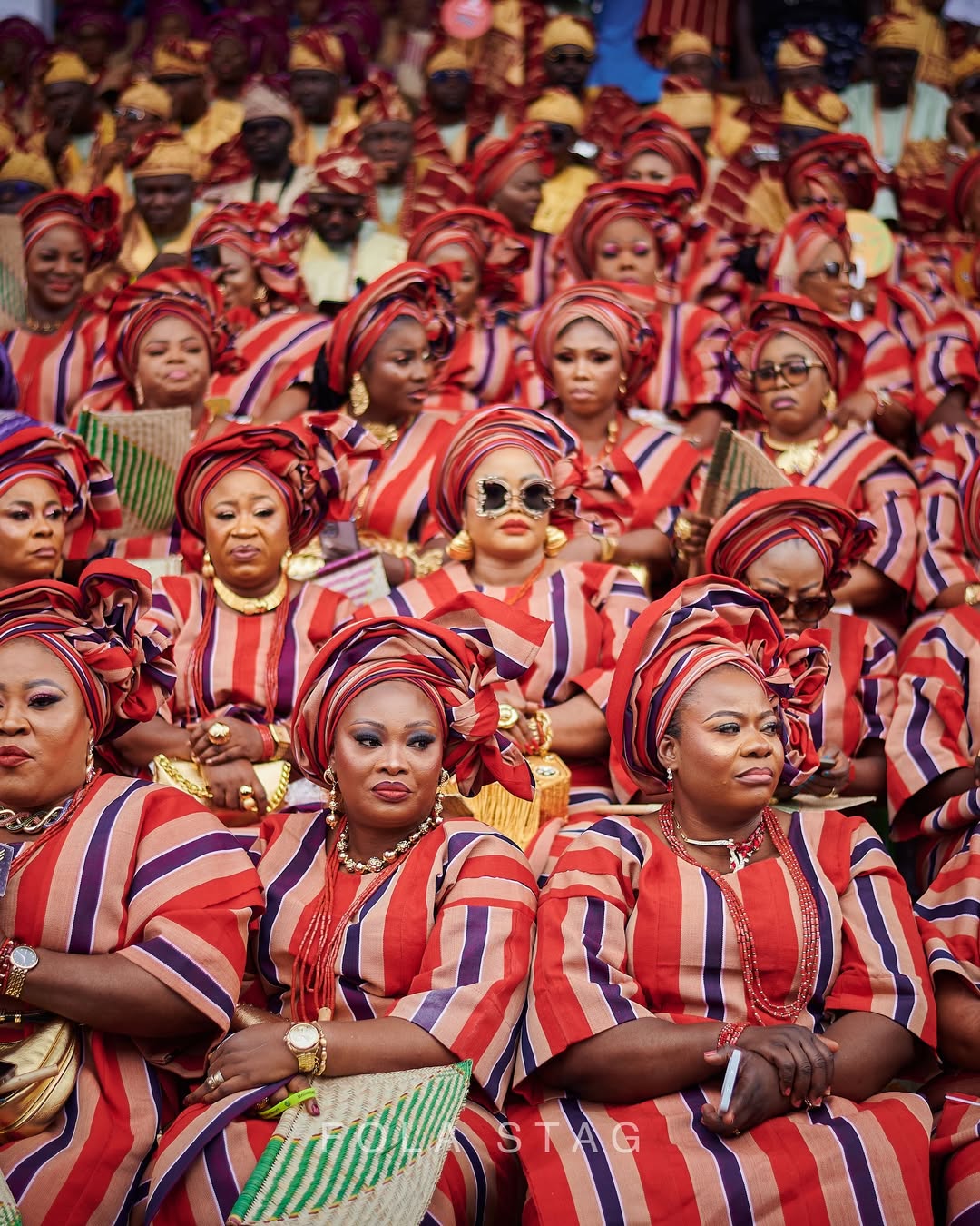 A large group of Nigerian women seated in rows at the Awujale Pavilion during Ojude Oba, uniformly dressed in crimson, navy blue, and cream striped traditional clothing and matching high-styled Gele headties.