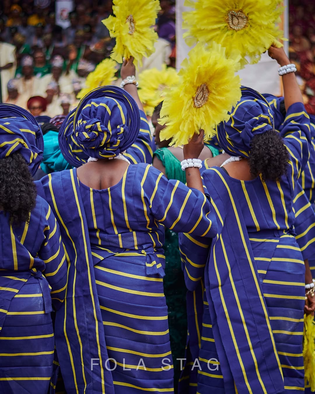 Rear view of a Yorùbá women's Regberegbe group at Ojude Oba wearing matching blue and yellow striped Asọ-Ọkè with structured Gele headgear and holding yellow feather fans.